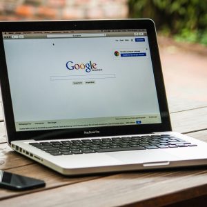 A MacBook Pro displaying Google Search on a wooden table outdoors, next to a smartphone.